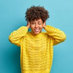 Horizontal shot of curly haired woman covers ears as hears very loud noise or music winks eye and clenches teeth ignores something noisy dressed in casual yellow jumper poses indoor over blue wall