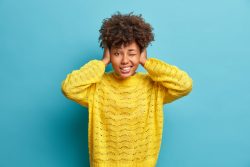Horizontal shot of curly haired woman covers ears as hears very loud noise or music winks eye and clenches teeth ignores something noisy dressed in casual yellow jumper poses indoor over blue wall