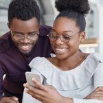 Happy African female in spectacles types message on modern smart phone while her boyfriend stands near her and looks at screen, have broad smile and white teeth. Dark young skinned woman and man