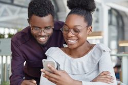 Happy African female in spectacles types message on modern smart phone while her boyfriend stands near her and looks at screen, have broad smile and white teeth. Dark young skinned woman and man
